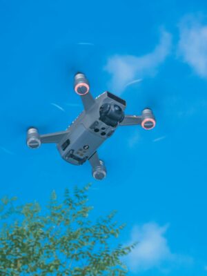 From below of modern UAV with photo camera and big propellers flying in sky above forest